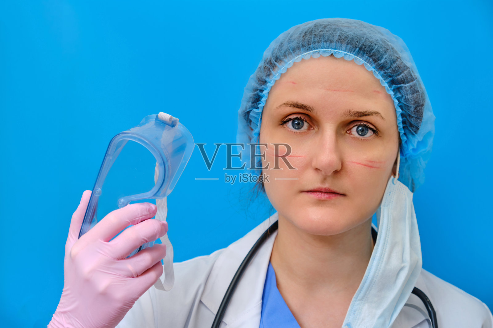 一位脸部受伤的医生在医院工作后摘下护目镜,蓝色背景。一个穿着白大褂戴着医用贝雷帽脸上有伤痕的护士。待在家里吧,冠状病毒。照片摄影图片