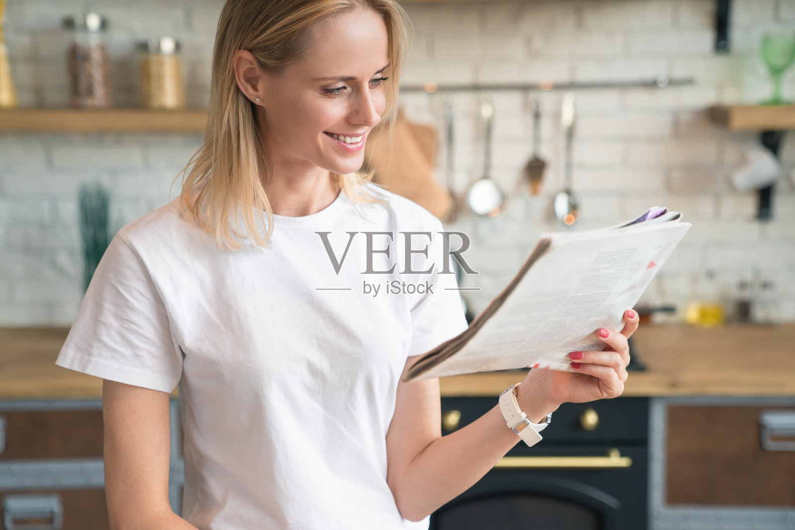 年轻美丽的女人正在阅读她的晨报,微笑着在厨房吃早餐。穿白衬衫照片摄影图片