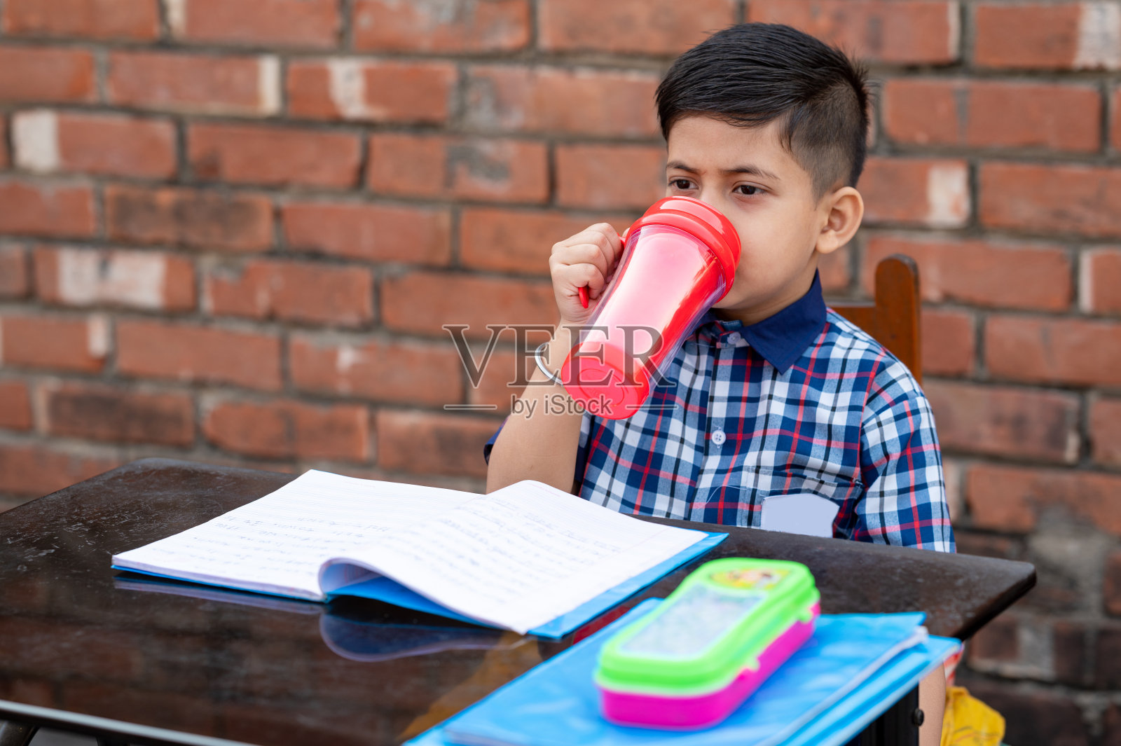 小学生坐在课桌上用红水瓶喝水。印度学校学生在教室里。照片摄影图片