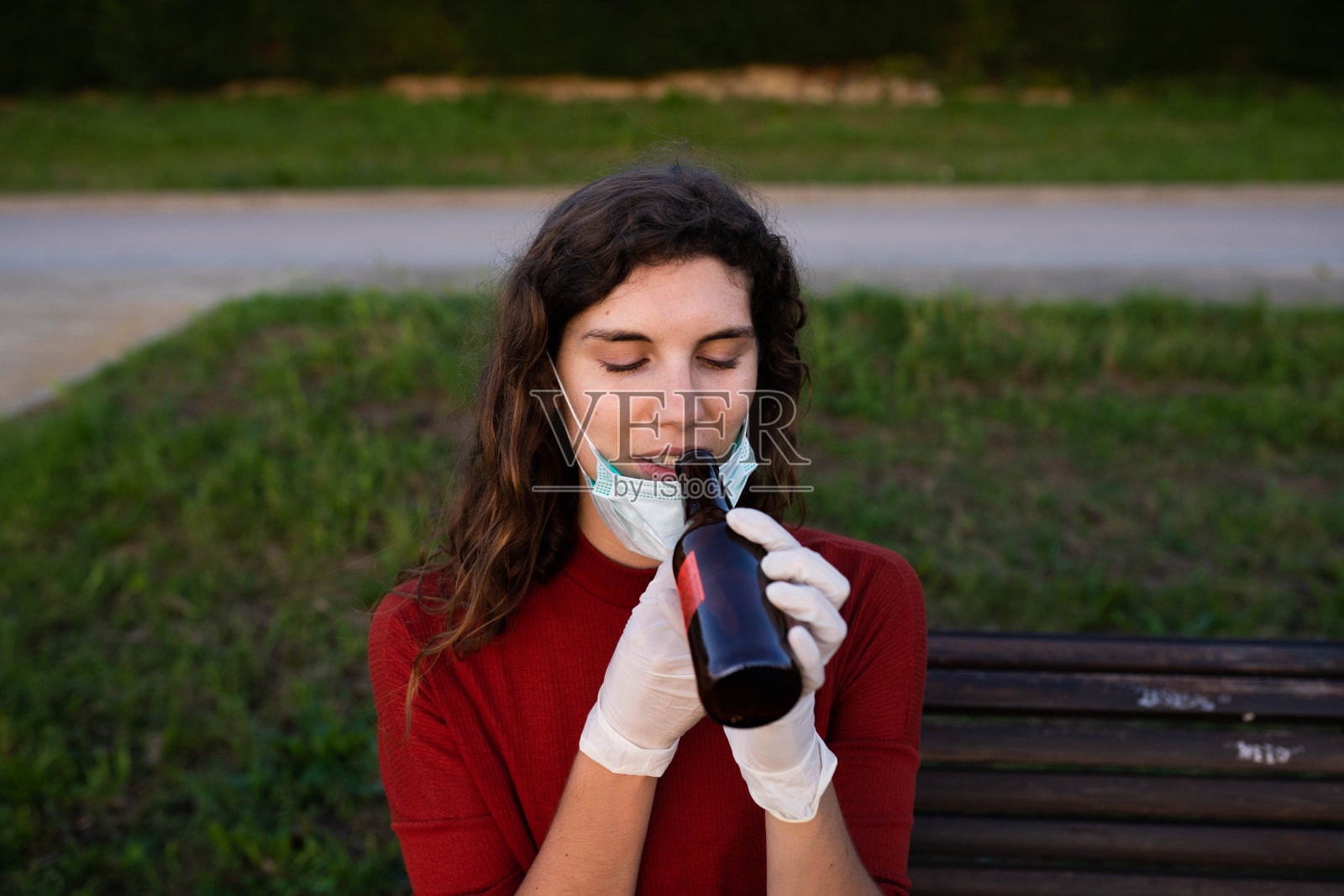 年轻女孩喝啤酒在公园的长凳上完全保护穿着面具和手套照片摄影图片
