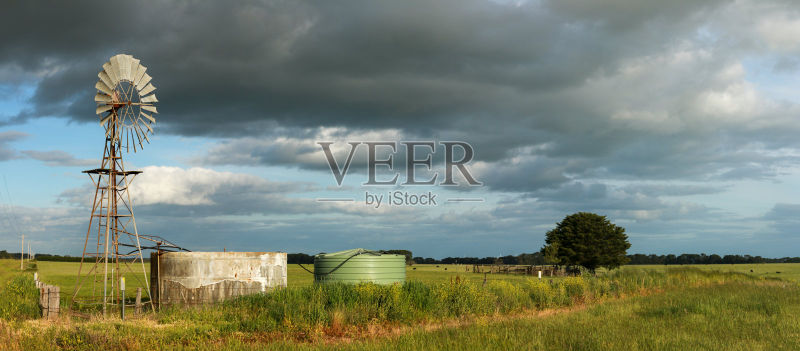 全景图片显示了在多云的天空下,长长的绿色农田与风力水泵和水泥罐在前景,汉密尔顿,维多利亚,澳大利亚农村照片摄影图片