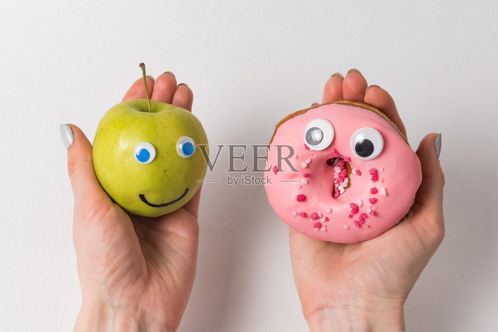 一只手拿着苹果微笑,另一只手拿着甜甜圈和谷歌眼睛。食物与有趣的面孔在白色的背景照片摄影图片