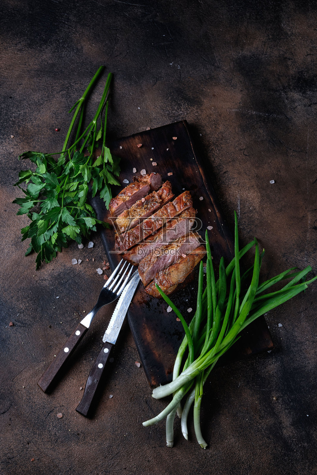 在切菜板和刀叉上放一片烤好的肉,配上欧芹和韭菜,在深色的背景上。俯视图照片摄影图片