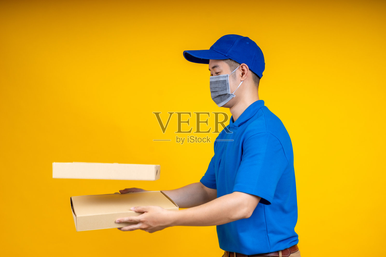 年轻的亚洲送货员拿着食物或披萨盒,并在黄色孤立的背景上显示应用手机。在家工作和交付概念。照片摄影图片