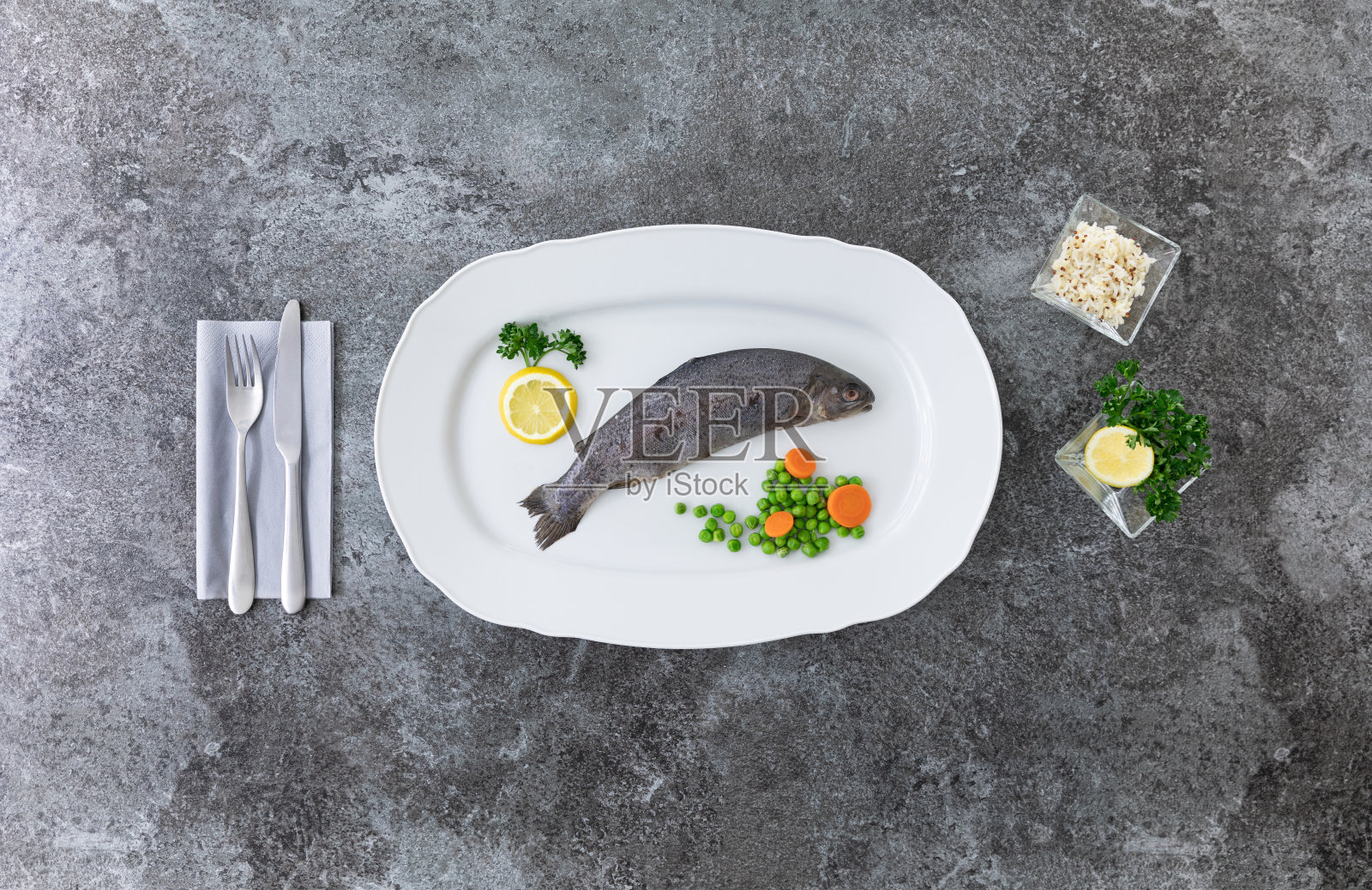 白色的盘子里放着一整条虹鳟鱼,灰色的大理石桌子上放着柠檬、欧芹、豌豆和胡萝卜,上面配有刀叉照片摄影图片