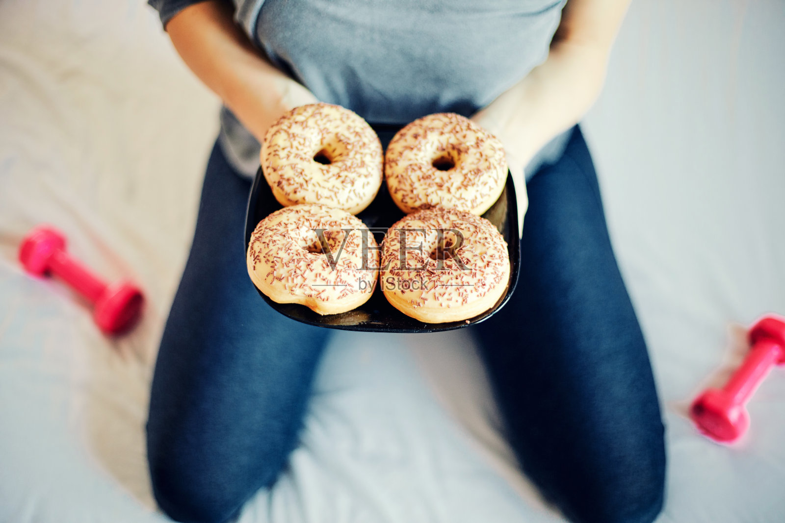 拿着巧克力甜甜圈和哑铃的漂亮女人。食品和运动概念。专注于甜甜圈。照片摄影图片