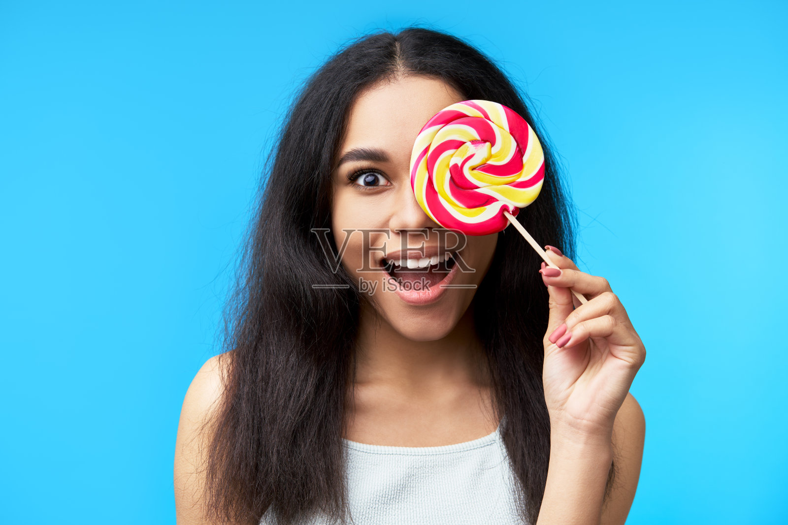 兴奋快乐的黑人妇女覆盖她的眼睛与彩色的棒棒糖孤立在蓝色的背景。照片摄影图片