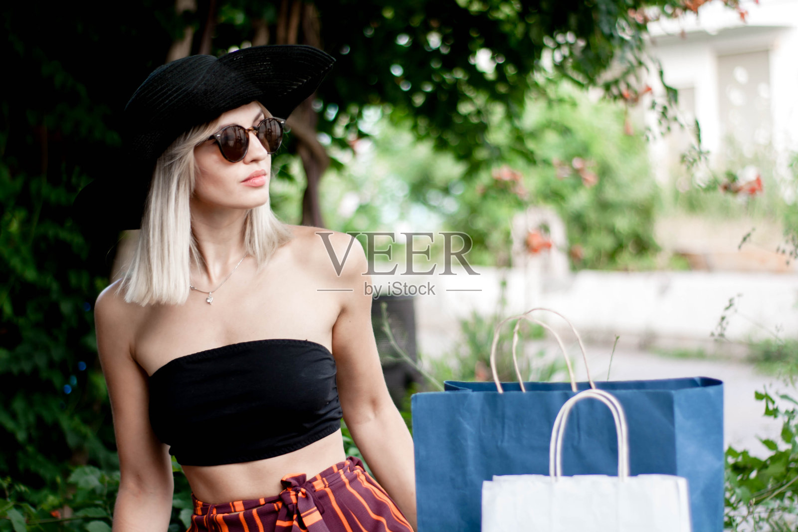 时髦的年轻女子戴着太阳镜,帽子和裤子在一条是时尚的穿着,去一个露天购物中心购物,并摆出彩色背景照片摄影图片