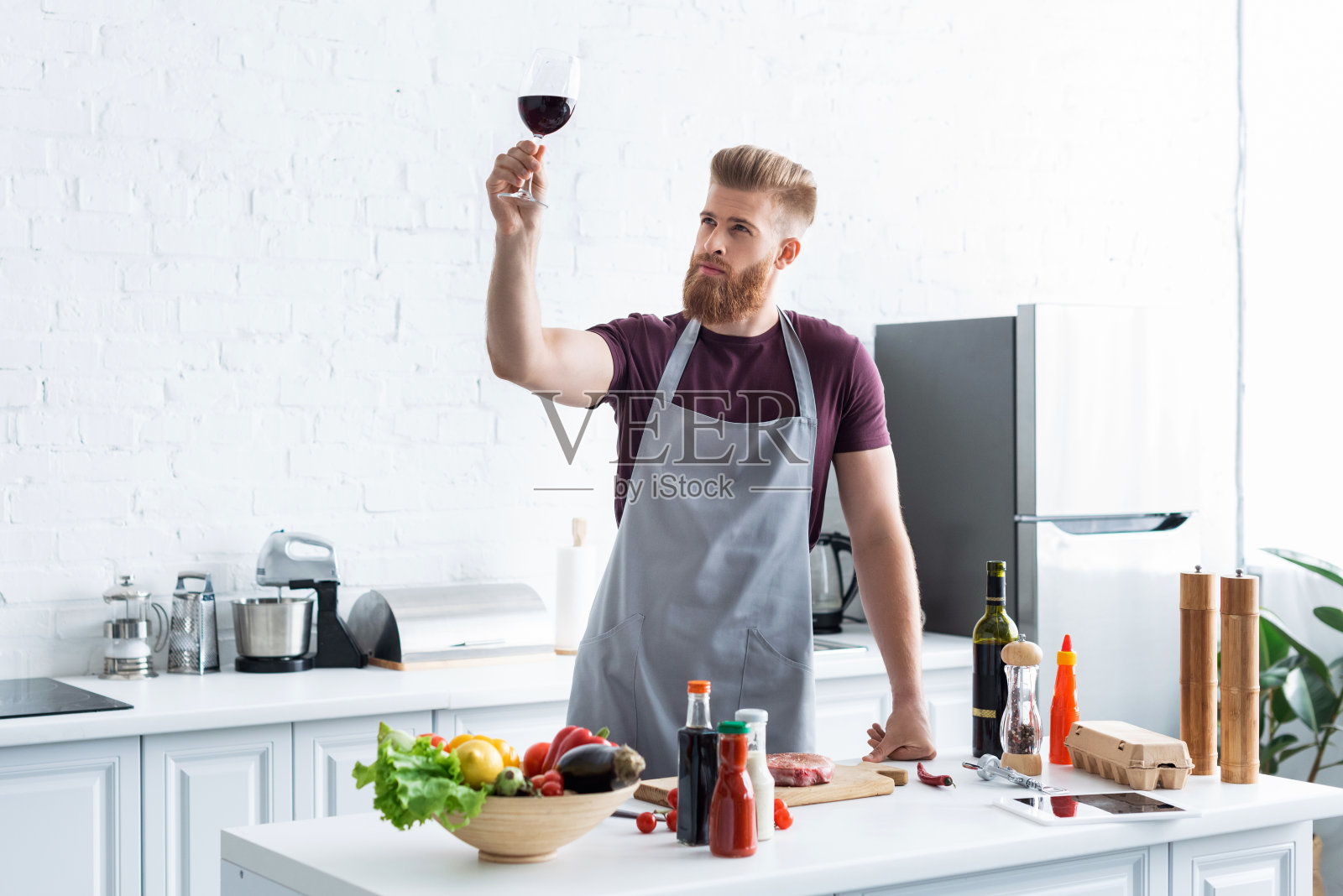 英俊的蓄着胡子的年轻人,穿着围裙,拿着一杯红酒在厨房做饭照片摄影图片