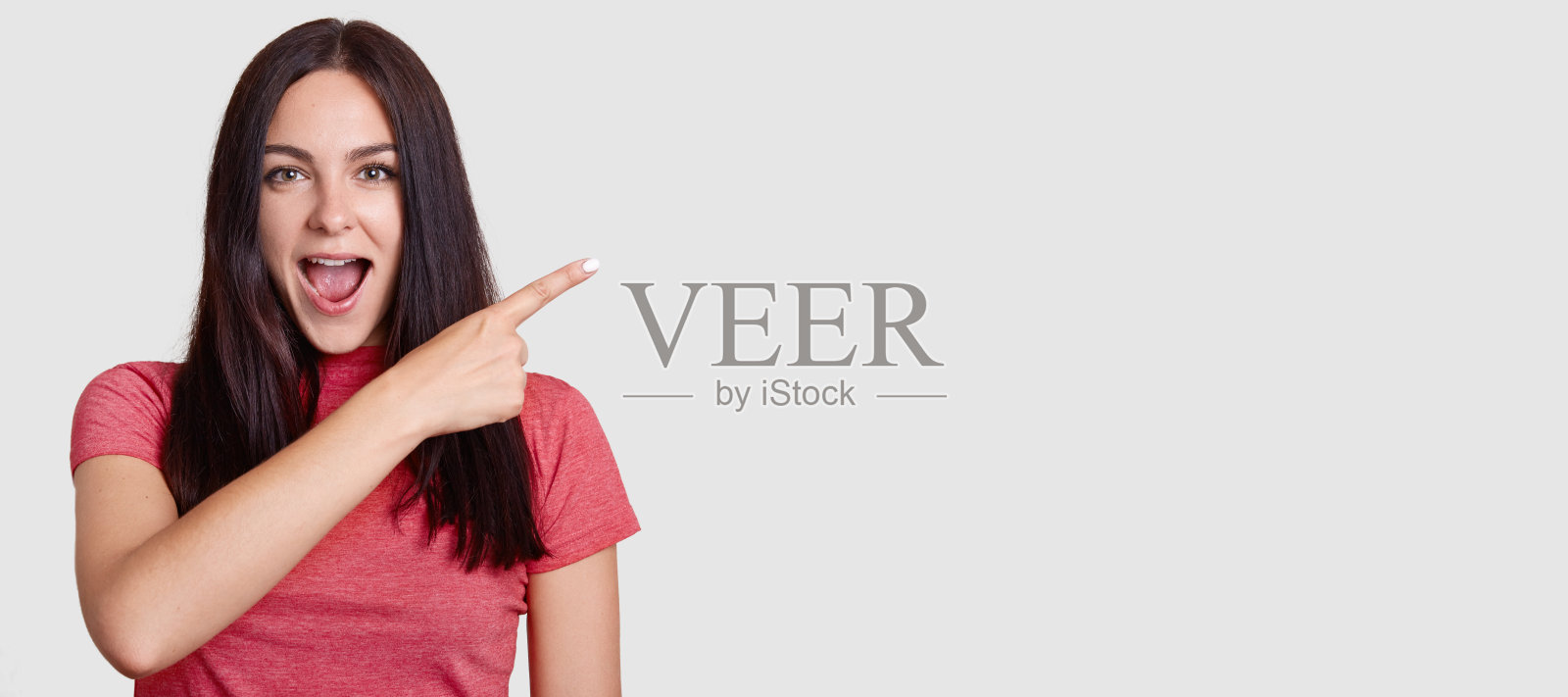 水平镜头惊讶的深色头发的女人,穿着粉红色t恤,用食指指着,显示你的晋升的自由空间,注意到一些难以置信的东西。看那里!照片摄影图片