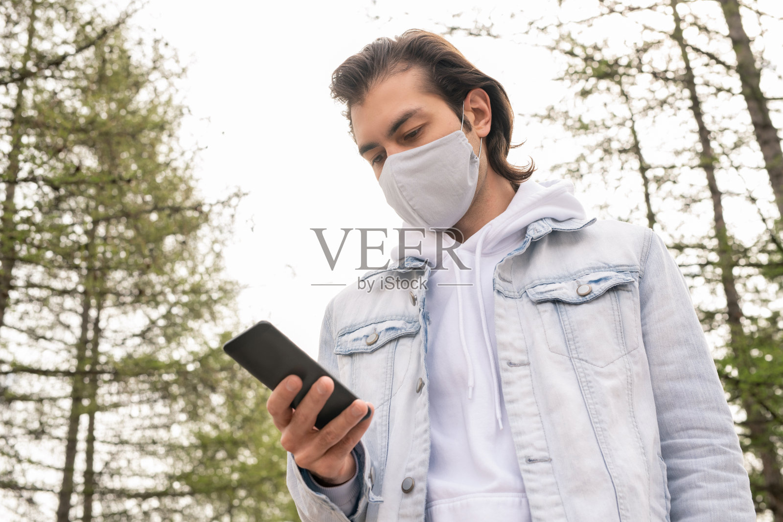 穿着牛仔夹克、帽衫和面罩的年轻人用智能手机滚动屏幕或发短信照片摄影图片