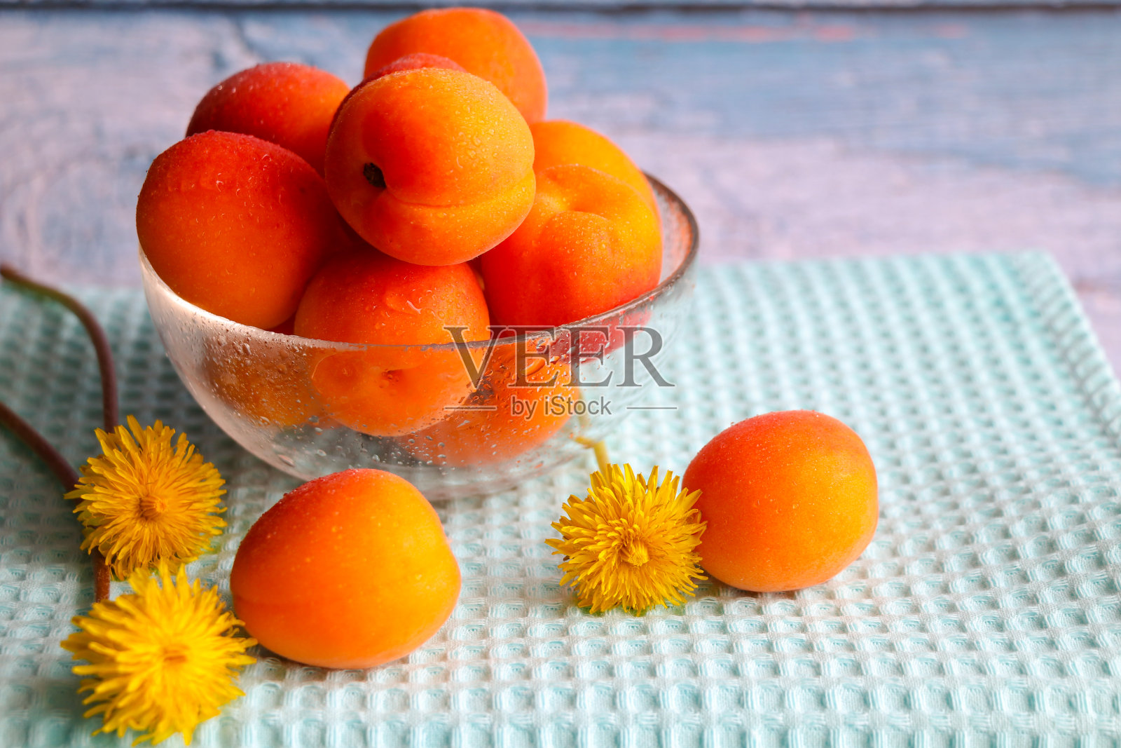 静物画上有新鲜的、成熟的杏子和蒲公英,背景是蓝色的木头。透明花瓶里放着鲜亮多汁的夏季水果。乡村风格。照片摄影图片