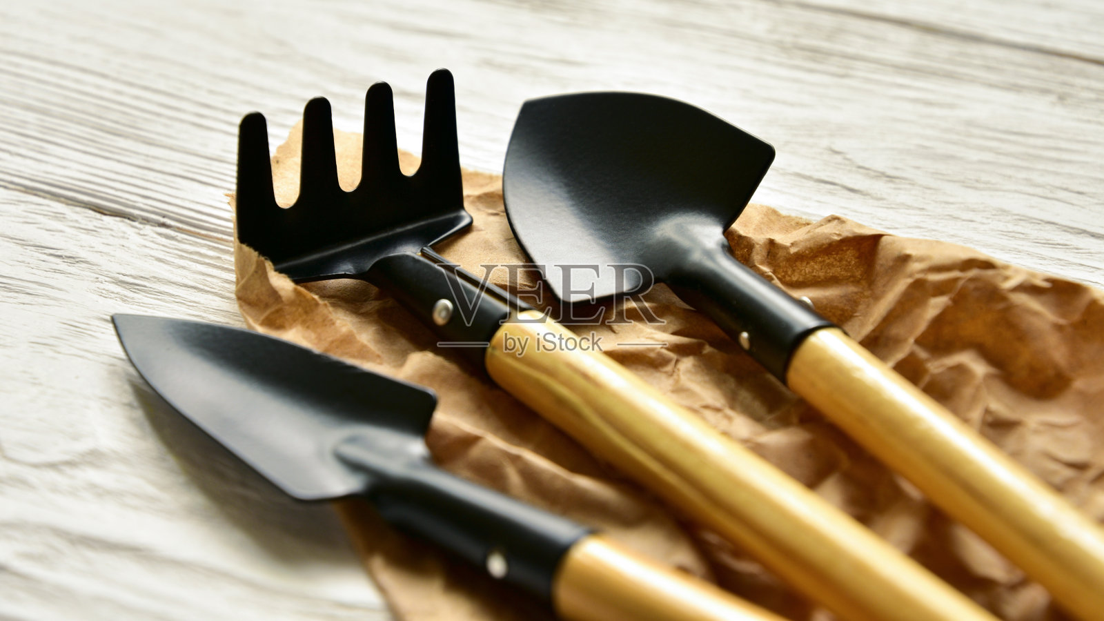 家庭园艺工具。两把小铲子和耙子放在牛皮纸和白色木质背景上。侧视图。特写镜头。照片摄影图片