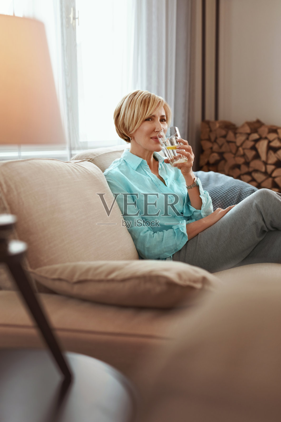 成熟的女人喝酒。中年女性穿着休闲服在家沙发上喝着柠檬汁。健康饮食的概念。照片摄影图片