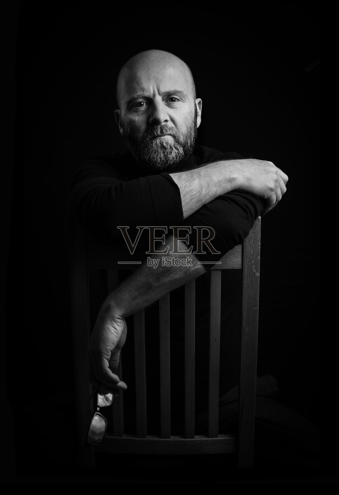 一个留着胡子的秃顶男人的简单画室肖像,胳膊随意地搭在椅背上,一只手里拿着一副眼镜,捕捉着光线。黑色背景,便于扩展复制。照片摄影图片