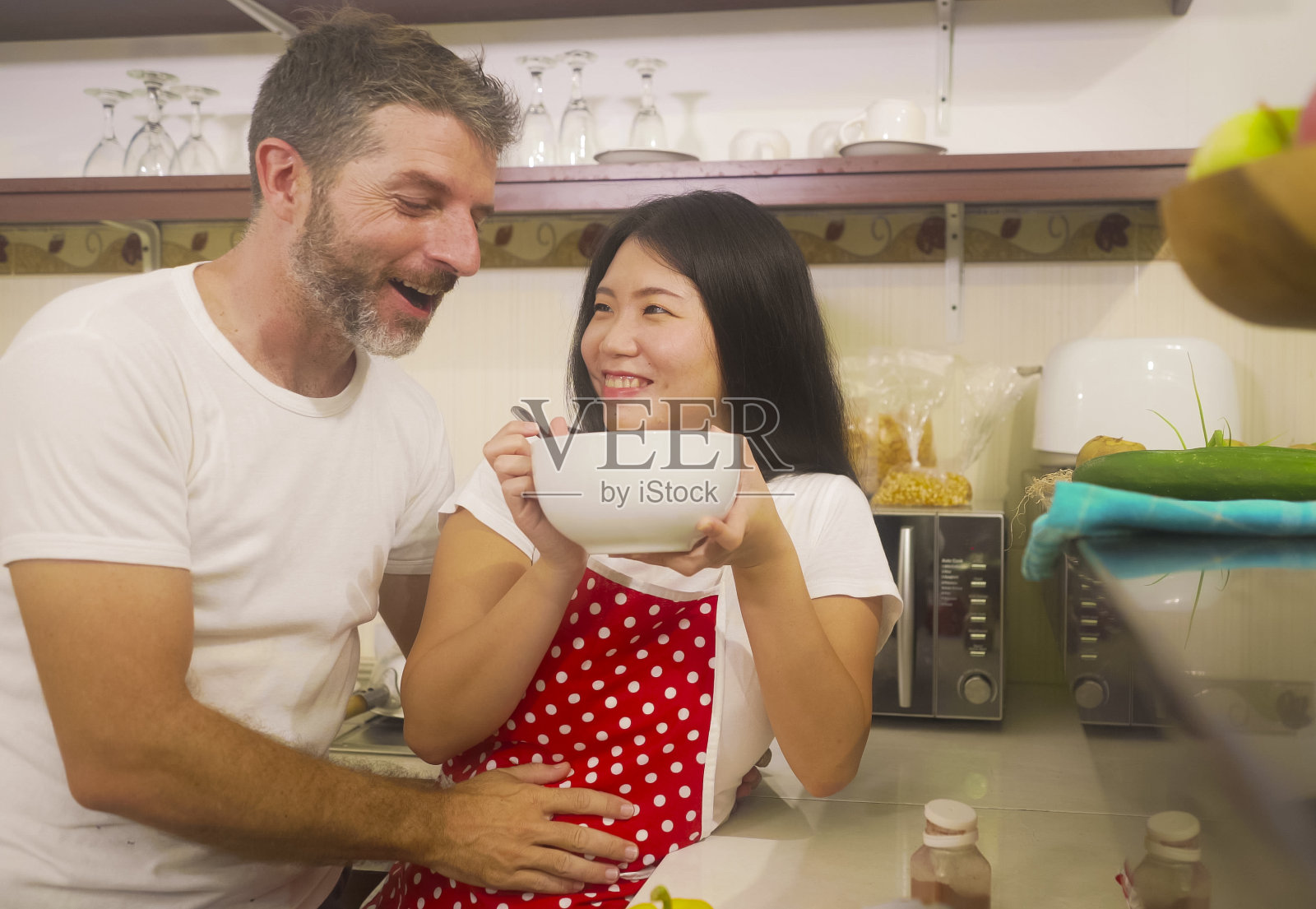 年轻美丽和幸福的混合民族夫妇在爱一起做饭在家里厨房的女人亚洲韩国人在红围裙和她的丈夫有乐趣的笑和享受照片摄影图片