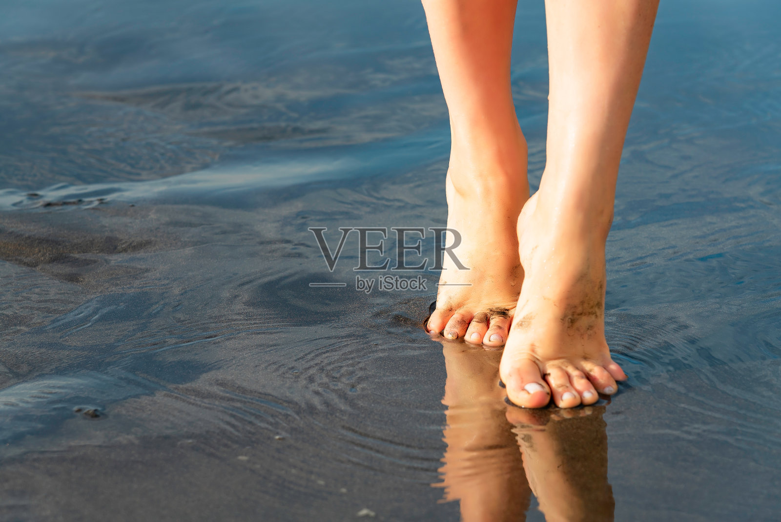 特写苗条的女人走在海边。湿漉漉的黑色沙滩上映出了女孩的双脚。照片摄影图片