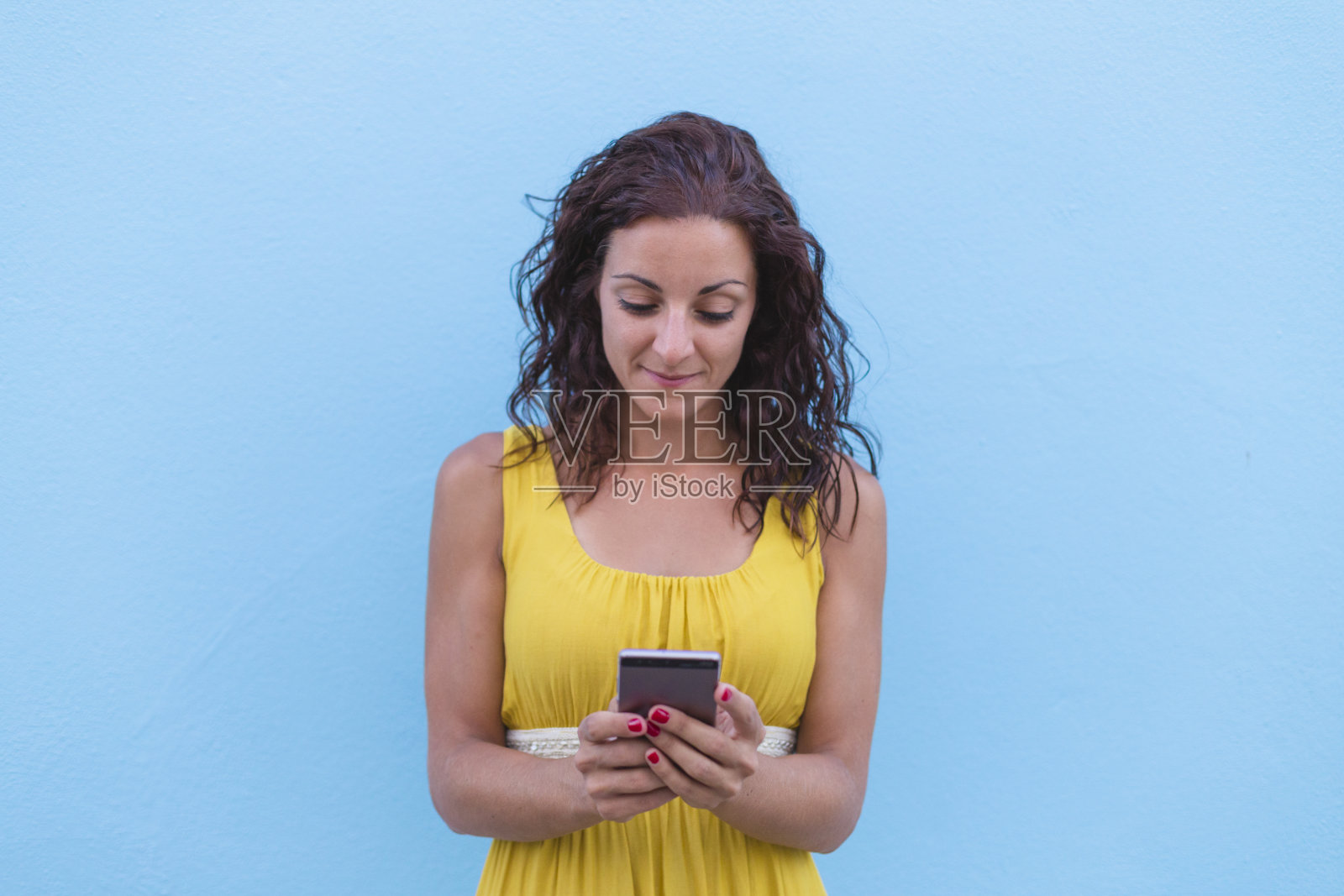 年轻漂亮的女人穿着黄色的衣服在一堵蓝色的墙上用手机。在户外。旅行的概念。城市照片摄影图片