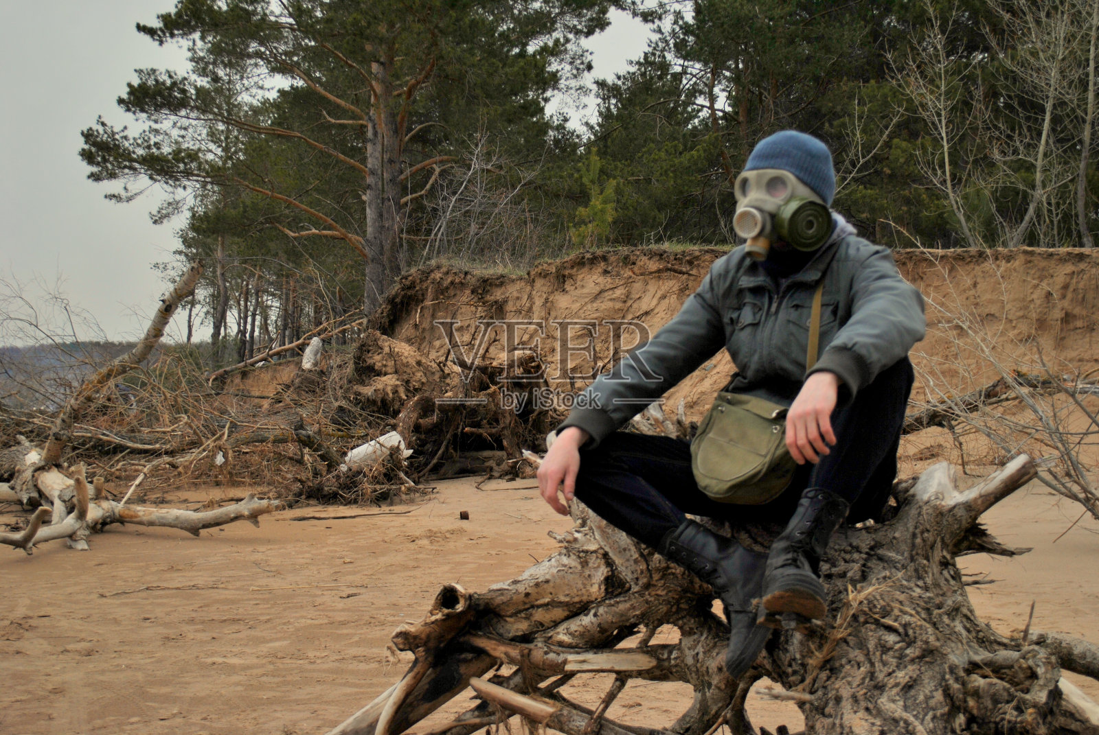 一个穿着灰色夹克、深色牛仔裤和防毒面具的男人坐在河边一棵枯死的树上,背景是灰色的夜空和松树林。照片摄影图片