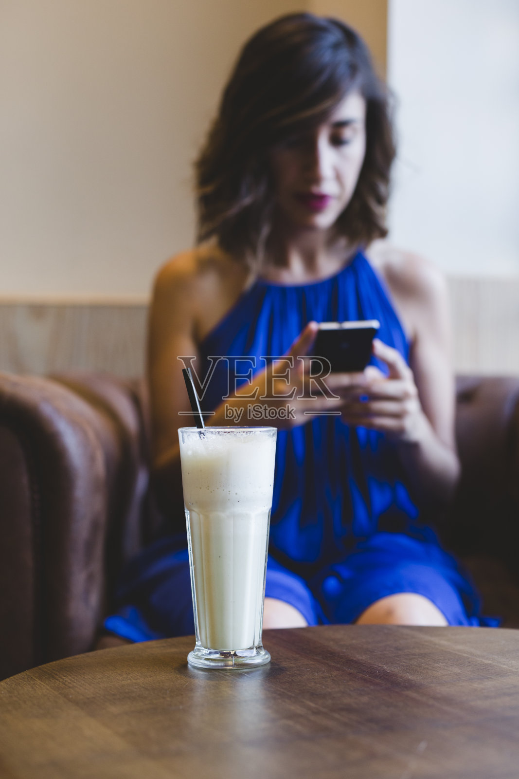 年轻美丽的女人享受一个冰沙饮料在一个咖啡馆和使用手机。休闲的蓝色裙子。现代生活在室内。有选择性的重点照片摄影图片
