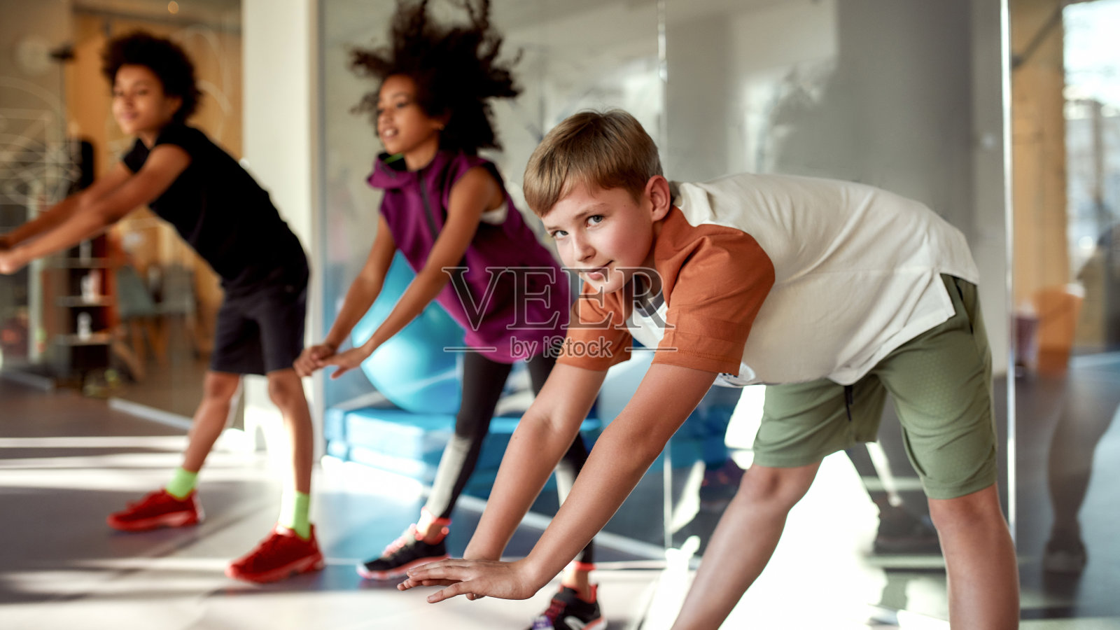 锻炼你的身体。一个男孩看着镜头,热身,和其他孩子一起在健身房锻炼。运动,健康的生活方式,积极的童年概念照片摄影图片