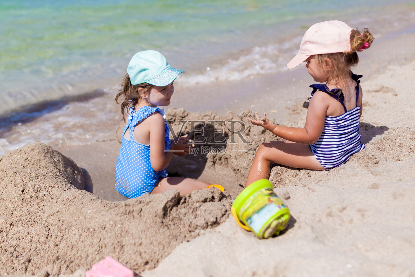 在一个阳光明媚的夏日,两个穿着泳衣和帽子的姐妹女孩在沙滩上玩玩具和沙子。有趣的海边家庭度假。孩子们在户外活动照片摄影图片