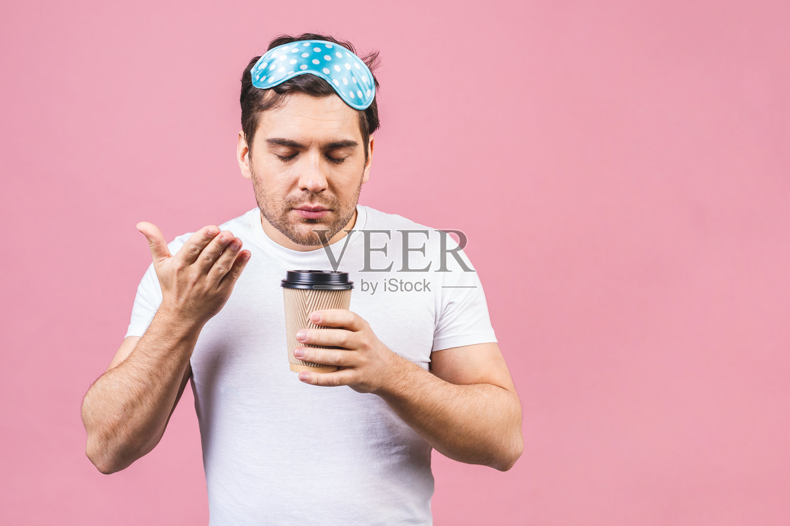这是一个安静的、安静的、昏昏欲睡的年轻人的肖像,他戴着蓝色的睡眠面具,喝着咖啡或茶。孤立的粉红色背景。照片摄影图片
