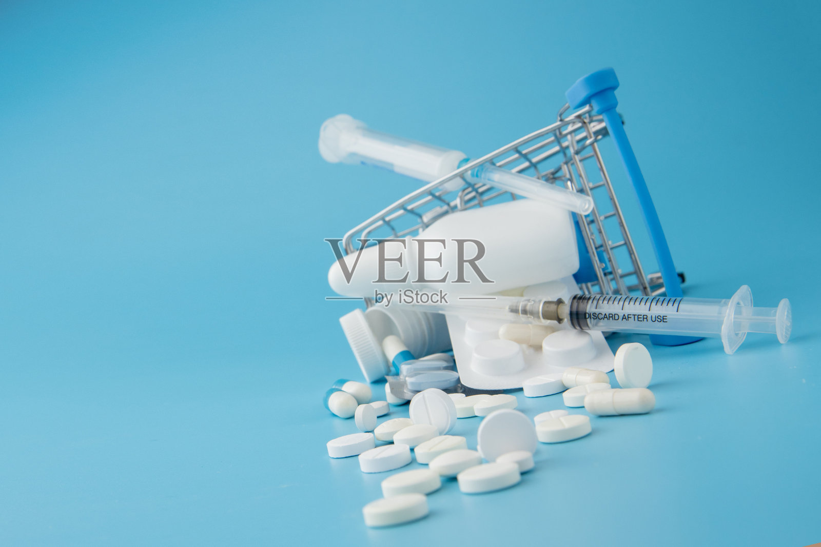 蓝色背景上散落着各种药片、药品、喷雾、瓶子、温度计、注射器和空的购物车。药房购物的概念照片摄影图片