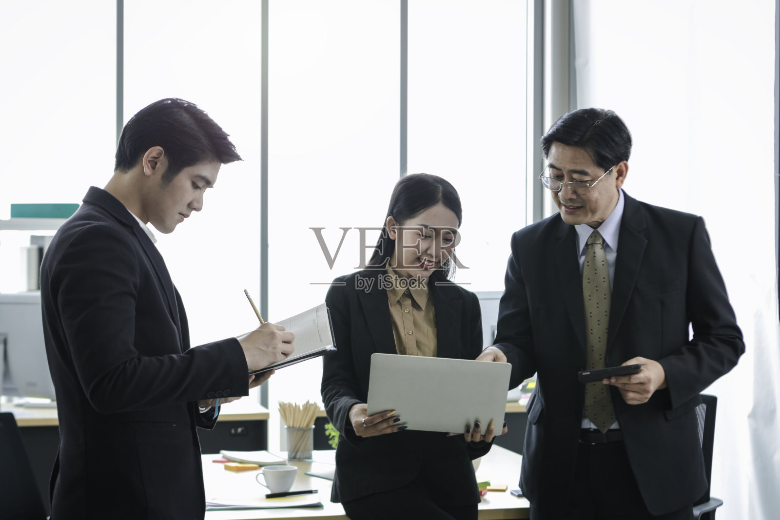 快乐的商务人士站在办公室一起讨论项目工作,商人和女商人一起使用笔记本电脑,而年轻人在他的笔记本上做笔记,人们在工作场所一起工作。照片摄影图片