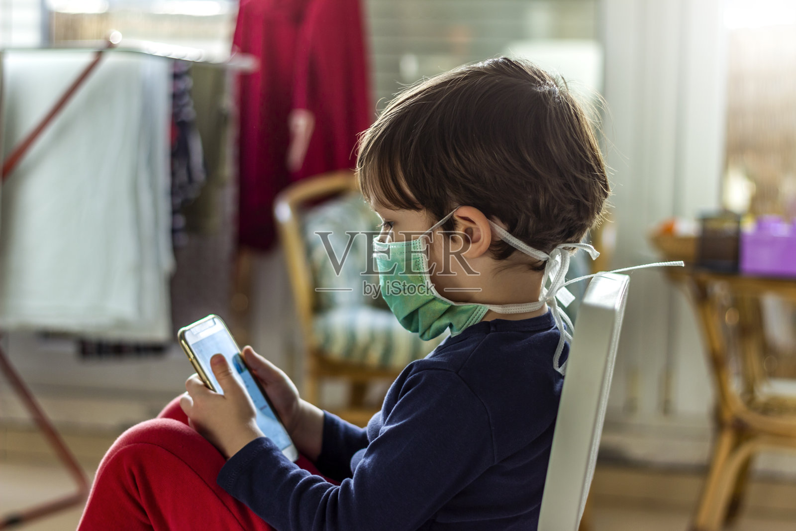 男孩戴着医用防护口罩玩智能手机游戏。照片摄影图片