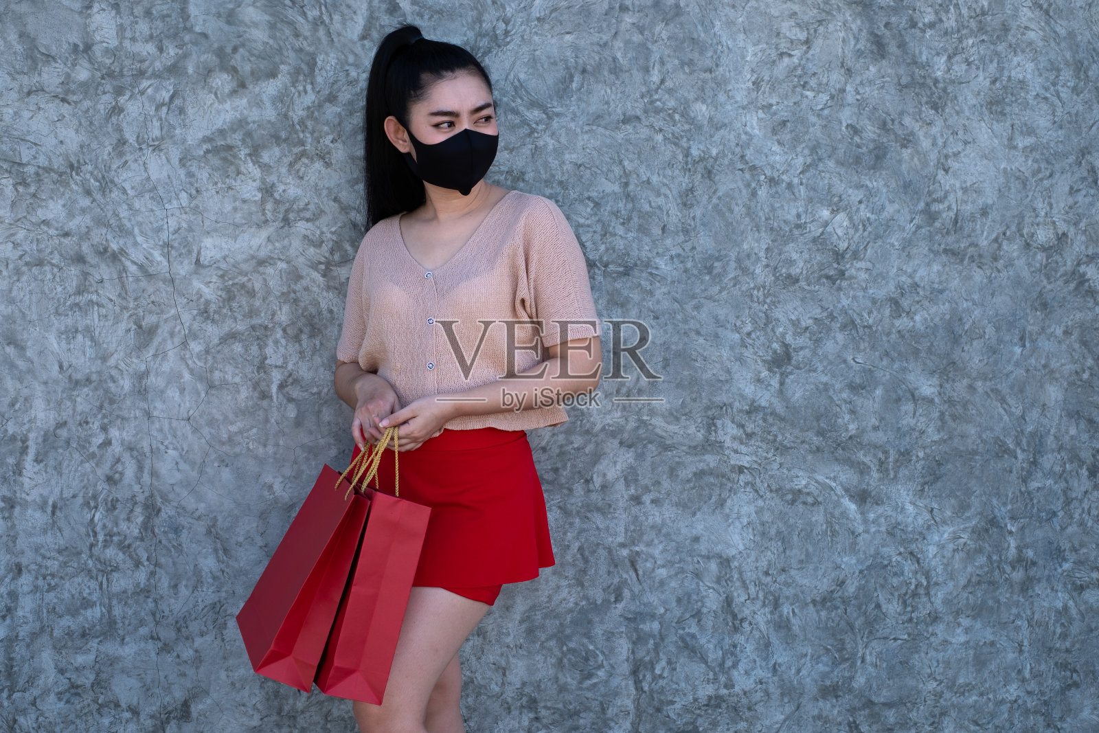 一个美丽的年轻亚洲女人的肖像戴着口罩,拿着购物袋在混凝土墙的背景,妇女穿粉红色的衣服和红色的裙子照片摄影图片