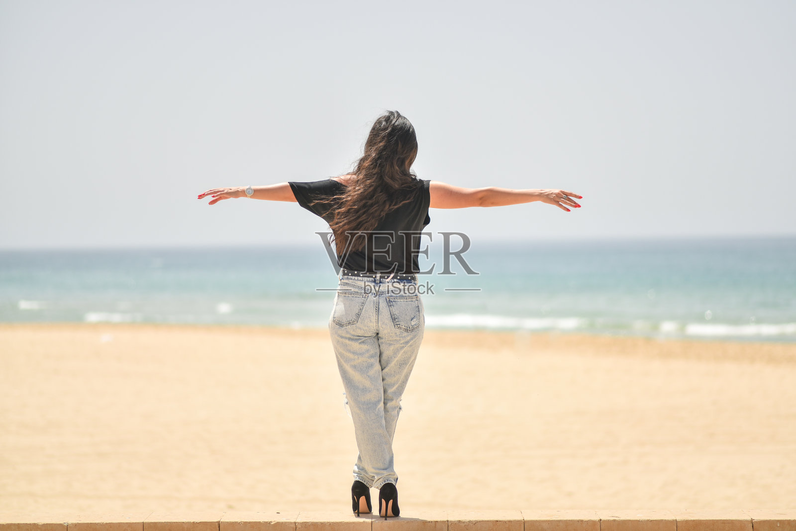 穿着黑色T恤、戴着时尚太阳镜和蓝色牛仔裤的年轻女子站在海滩上,看着大海照片摄影图片
