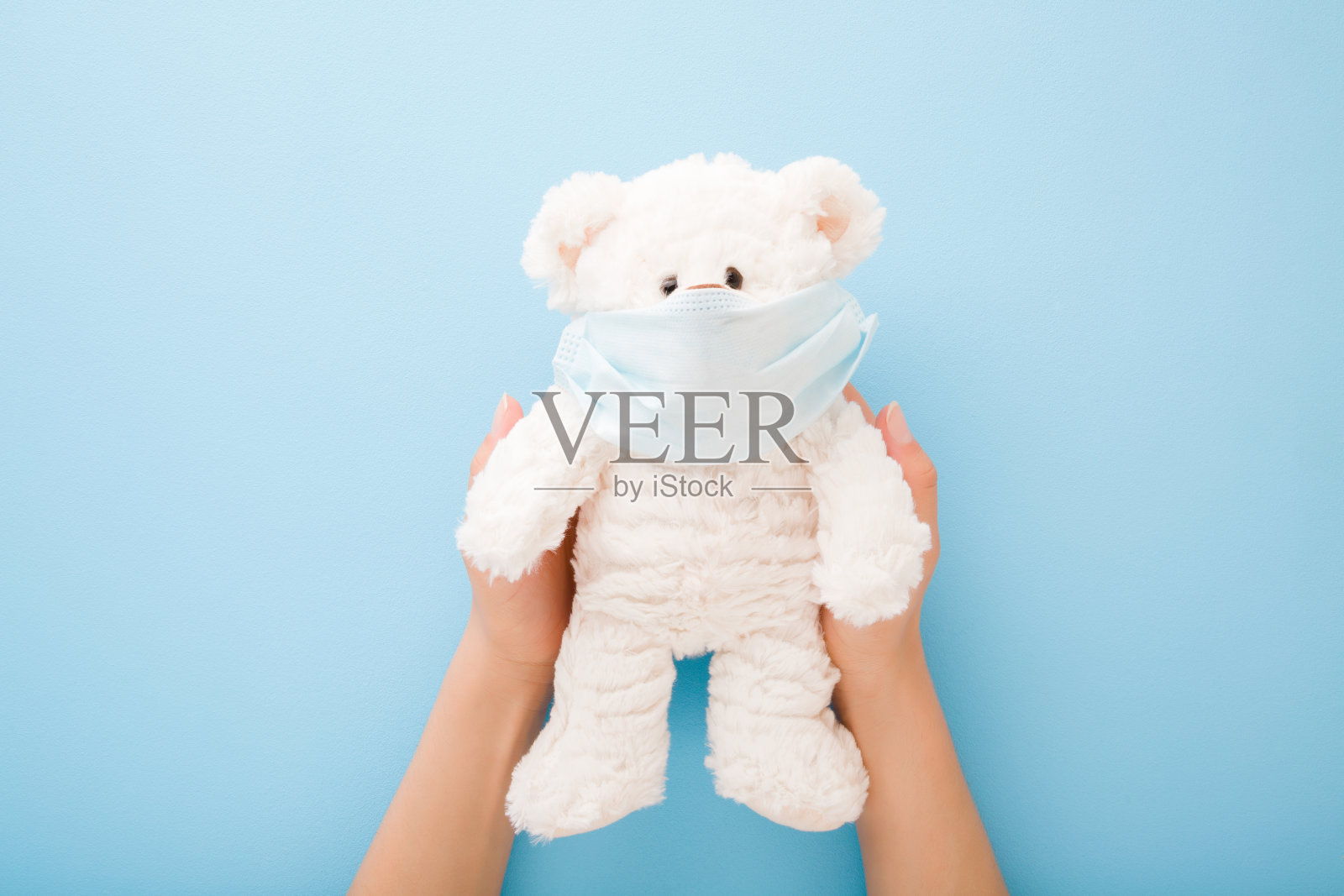 一名年轻女子双手捧着白色泰迪熊,戴着口罩捂住嘴和鼻子,桌子的背景是淡蓝色。第一次保护在危险的病毒时期。儿童医疗保健。前视图。照片摄影图片