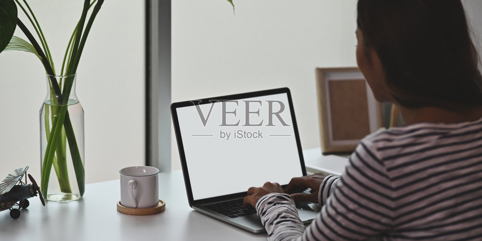 有创意的女人用白色空白屏幕的电脑笔记本电脑放在白色的办公桌上,周围是咖啡杯,花瓶里的植物,空的相框。照片摄影图片