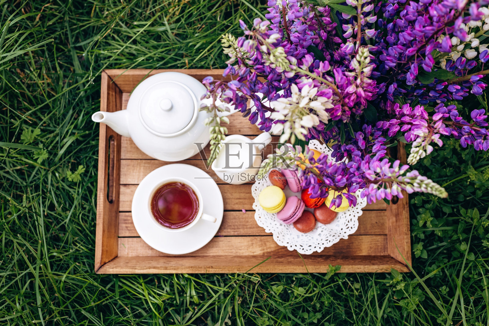在新鲜空气中享受一顿舒适的早餐。一个插着羽扇豆花的花瓶,一个茶壶,一杯咖啡茶,一个木托盘上放着杏仁饼干的蛋糕。母亲节或妇女节。软选择聚焦。照片摄影图片