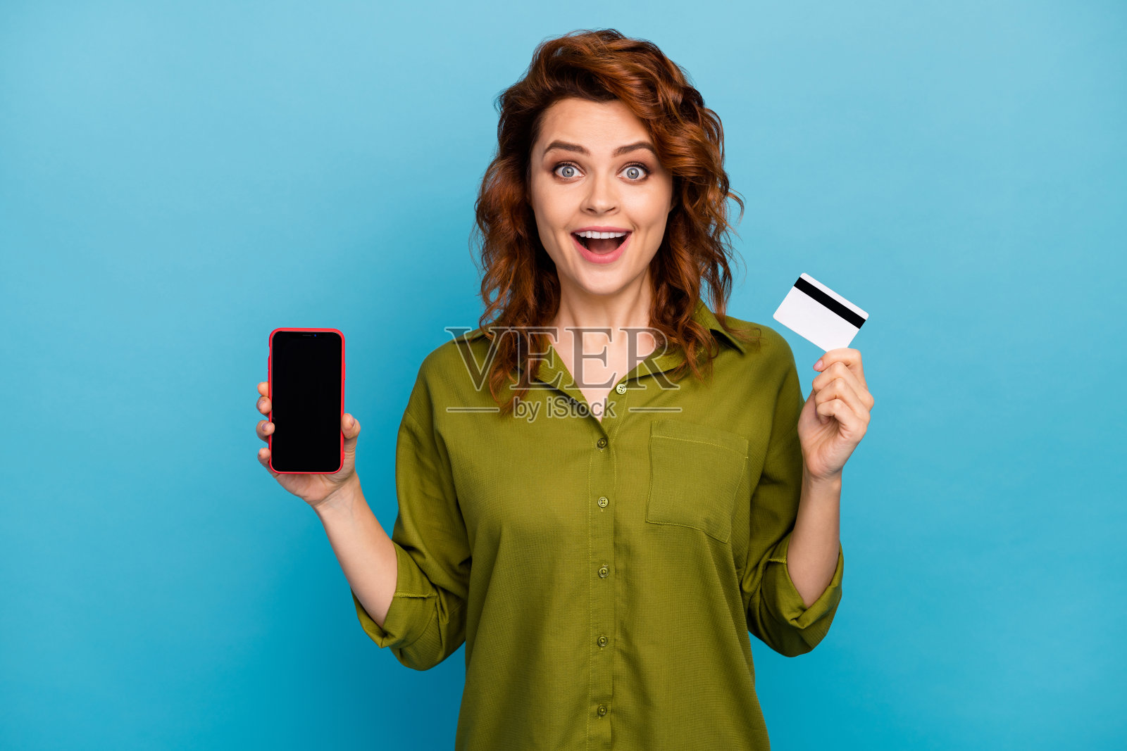 疯狂的红头发女人的肖像,拿着手机,印象深刻的支付信用卡,享受尖叫,哇哦,我的天,穿绿色的衬衫孤立在蓝色的背景照片摄影图片