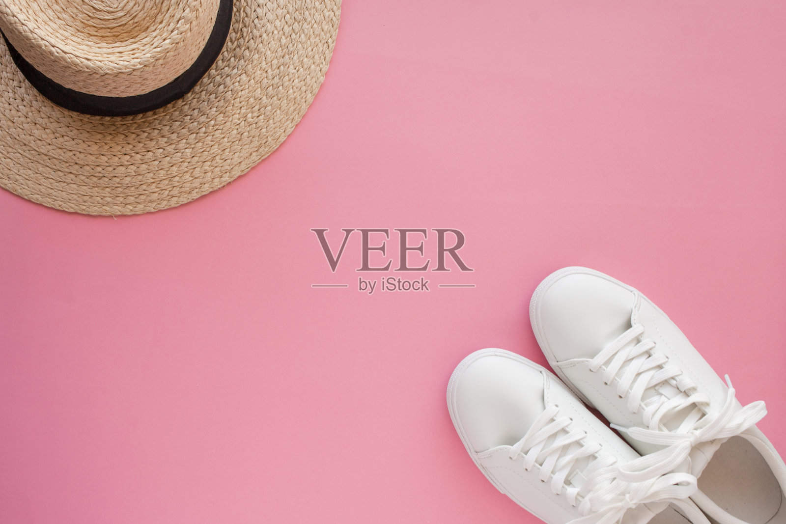 白色运动鞋躺在一顶草帽前的粉红色背景上。照片摄影图片
