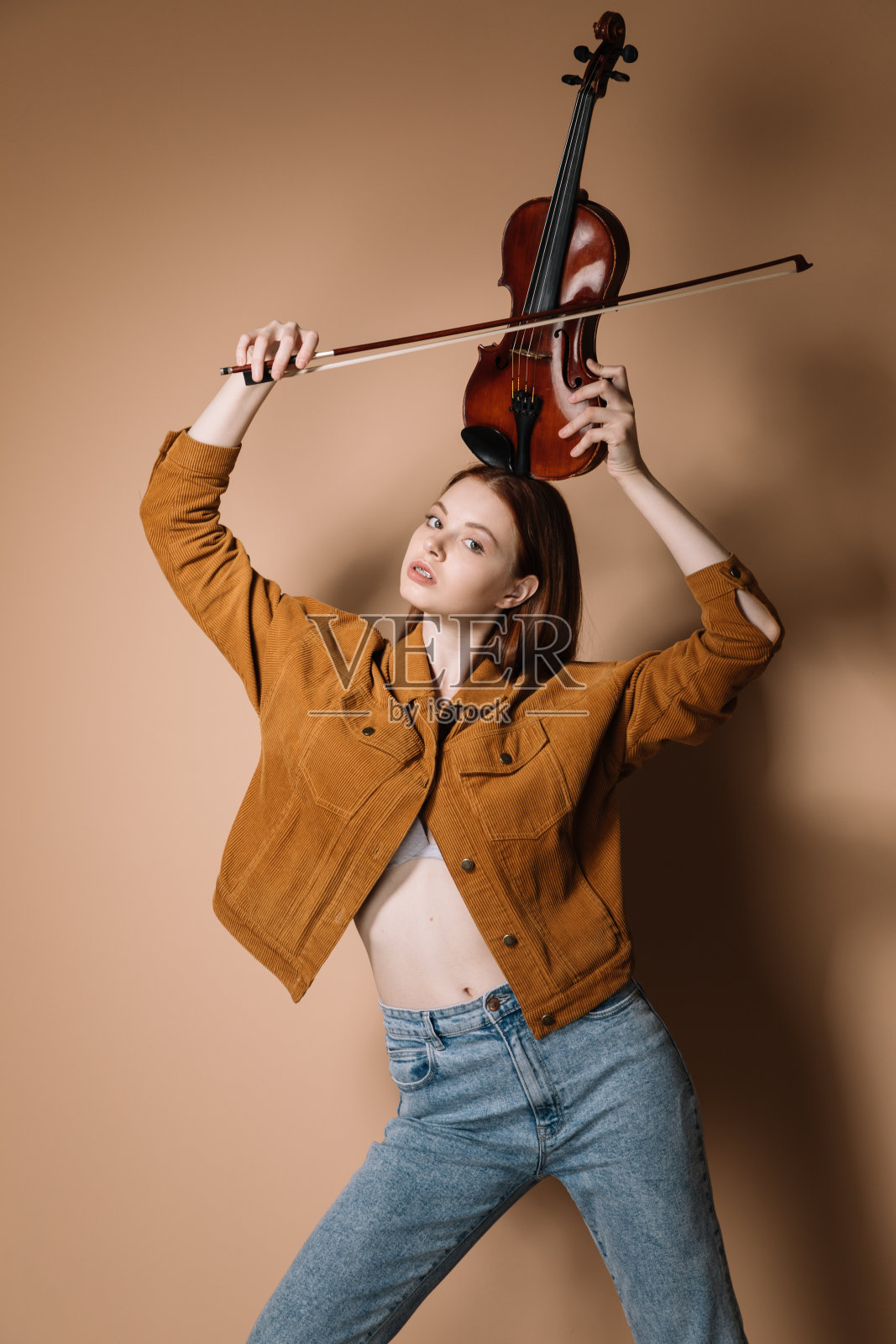 一个穿着牛仔裤和棕色夹克的女孩在米色的背景下拉着小提琴照片摄影图片