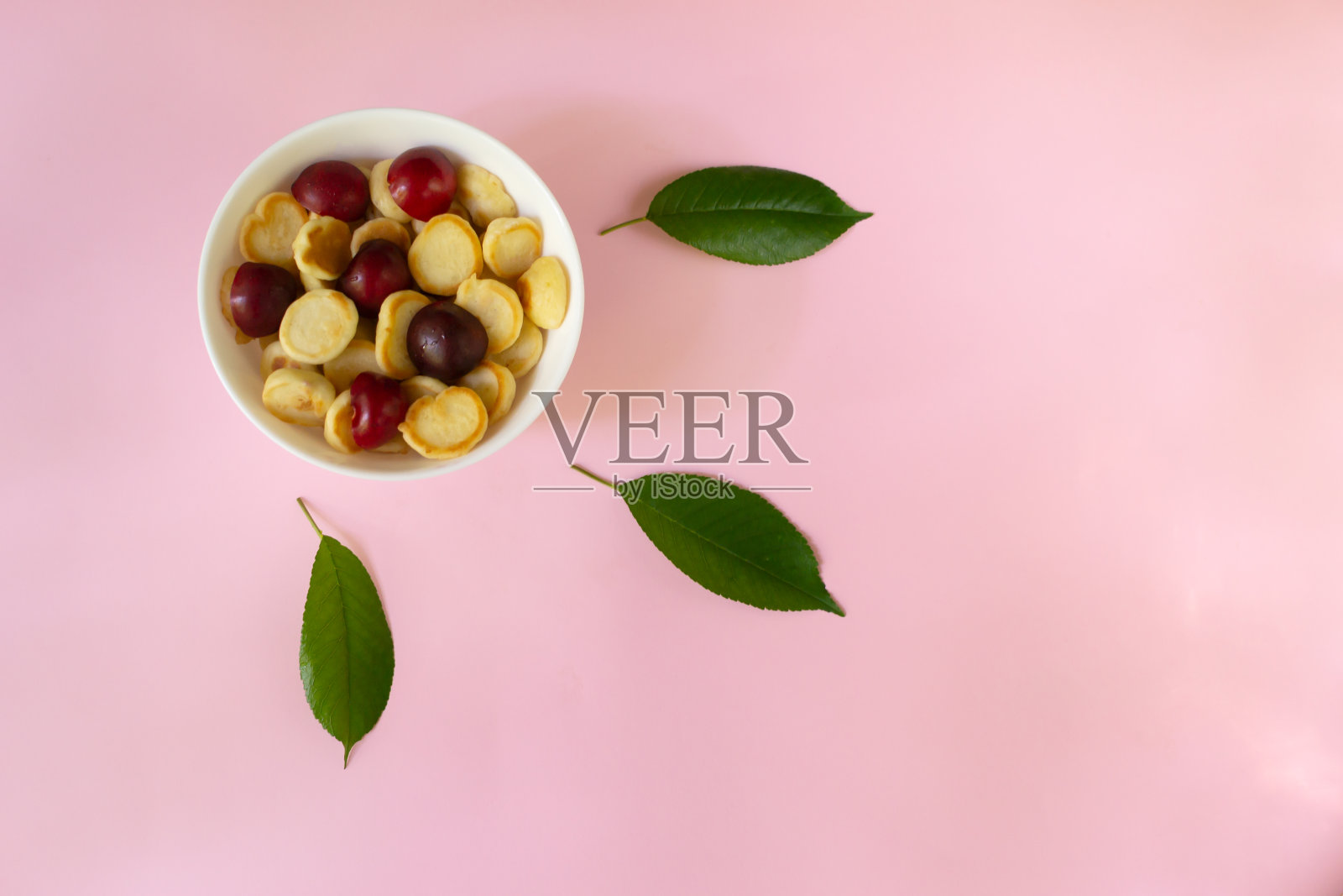 白色的碗,上面有一个小薄饼,粉红色的背景上有甜樱桃和绿叶。前视图。照片摄影图片