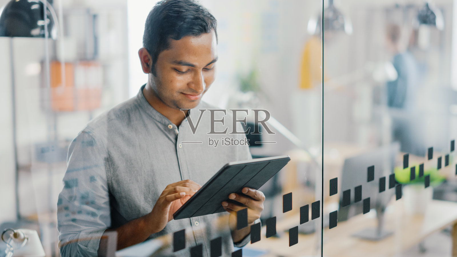 英俊的印度职业男子的肖像使用触摸屏数字平板电脑,写重要的电子邮件,迷人的微笑。在明亮多元的办公室工作的成功人士。照片摄影图片