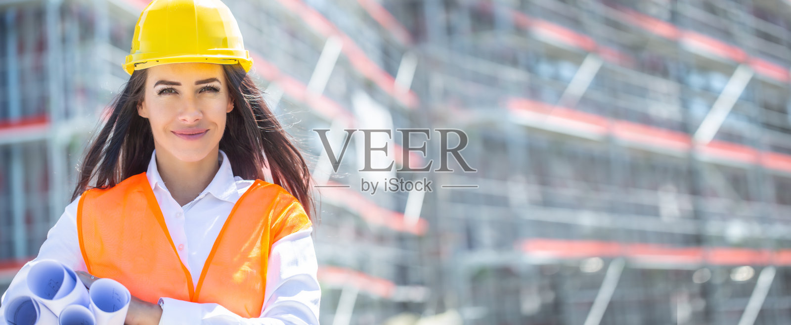 一名身穿橙色安全背心、头戴黄色头盔的女建筑师在未完工的大楼前,腋下夹着卷好的施工计划。照片摄影图片