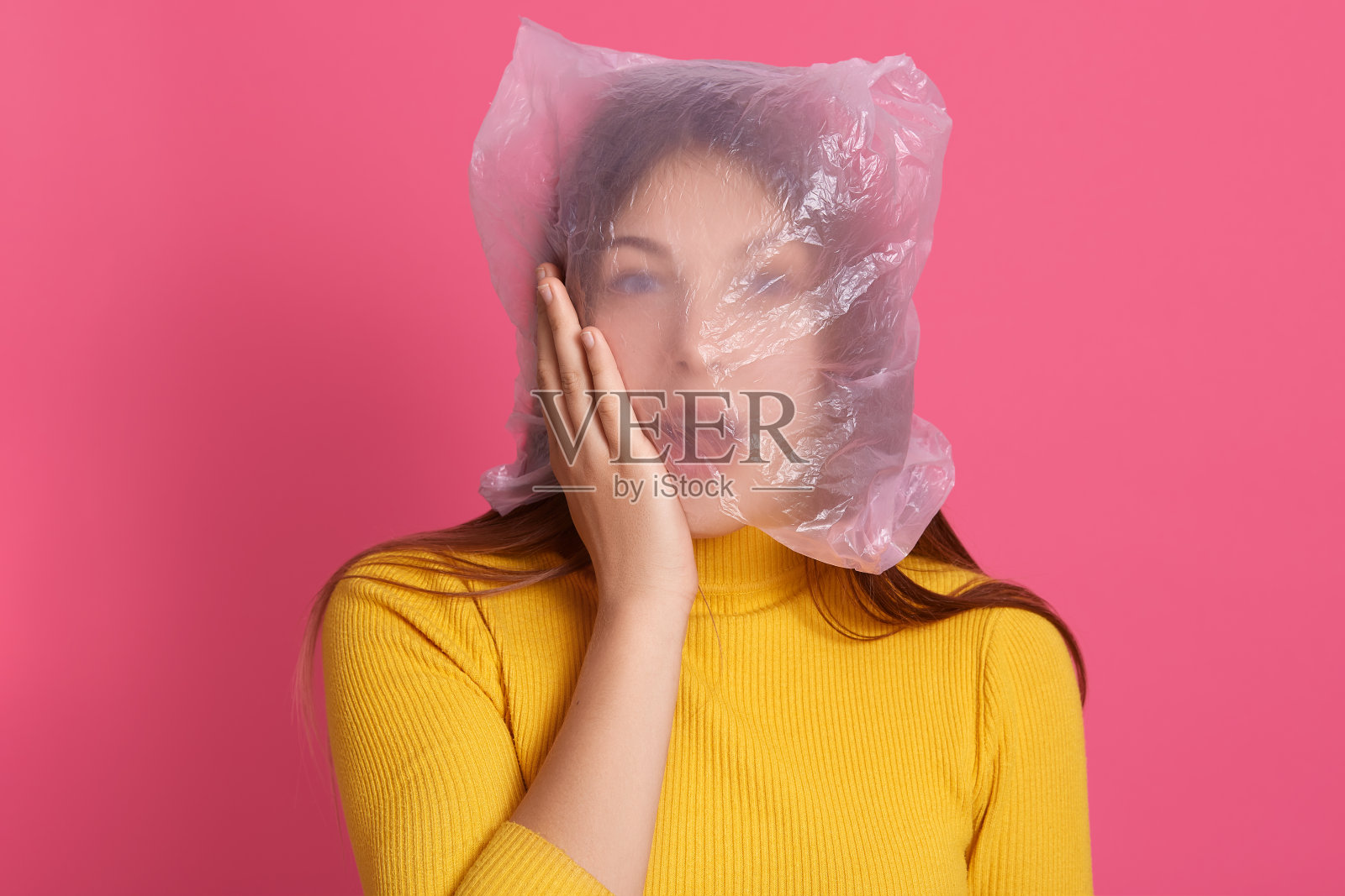 照片中,一名惊讶的女性张着嘴,头戴塑料袋,手掌放在脸颊上,穿着黄色休闲服,看起来很震惊。照片摄影图片
