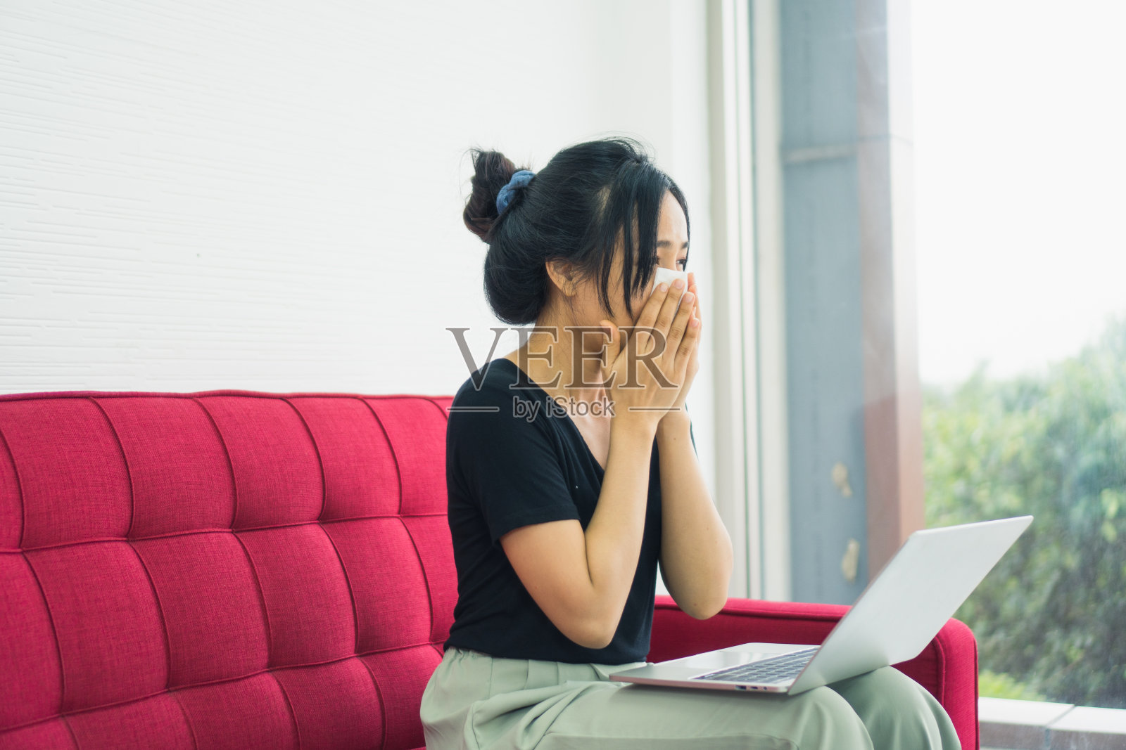 年轻漂亮的亚洲女人得了鼻过敏症,感冒打喷嚏的鼻子坐在客厅的红色沙发上伤心的感觉生病的工作形式的家。冠状病毒、COVID-19大流行新常态家庭生活概念照片摄影图片