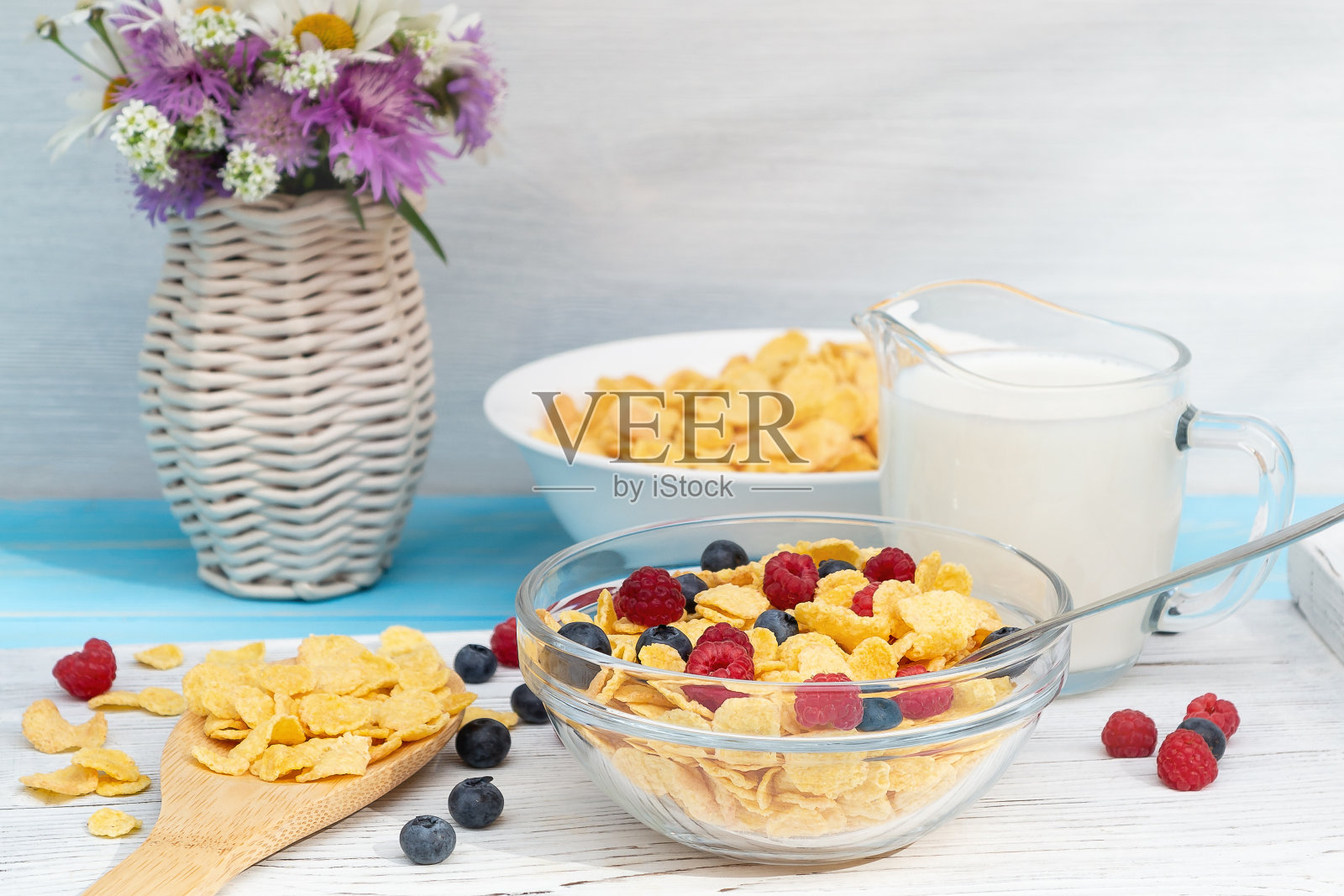 健康的夏季早餐-麦片,牛奶和新鲜的浆果碗在桌子上。浅色木质背景,花瓶里插着野花。健康饮食的概念照片摄影图片