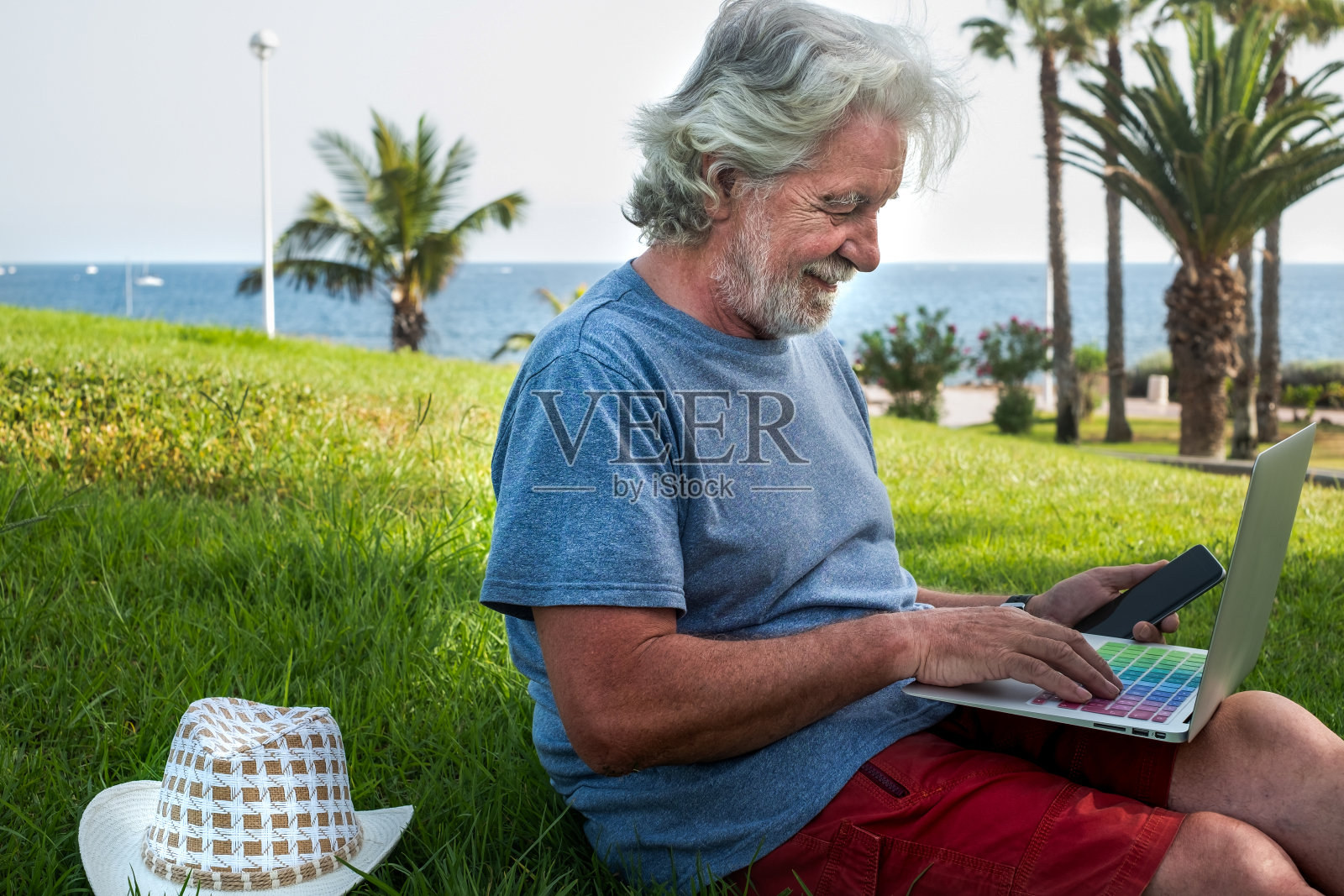 一个白胡子白头发的老人坐在草地上,用笔记本电脑和书工作和学习。背景是蓝色的海水照片摄影图片