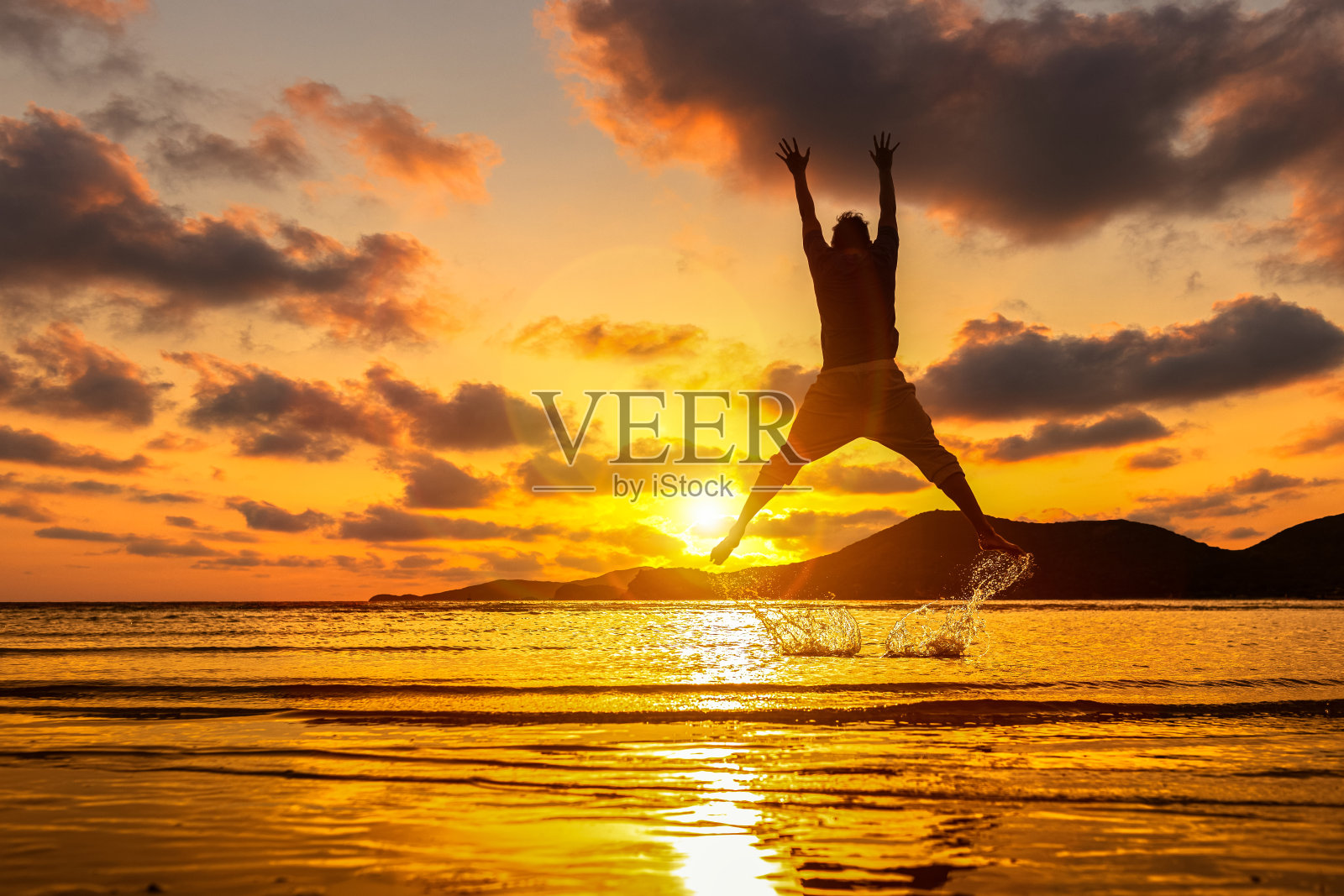 快乐的男人在海滩上跳跃,溅起水花。山,海,水和美丽的云是风景。照片摄影图片