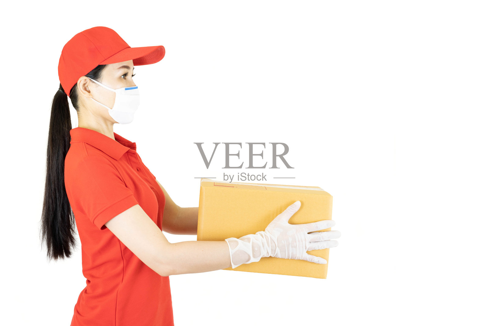 身穿红色制服的送货女工被隔离在白色背景上。快递员在防护口罩和医疗手套,帽子,t恤作为经销商拿着纸板箱。收到包裹。复制空间广告照片摄影图片