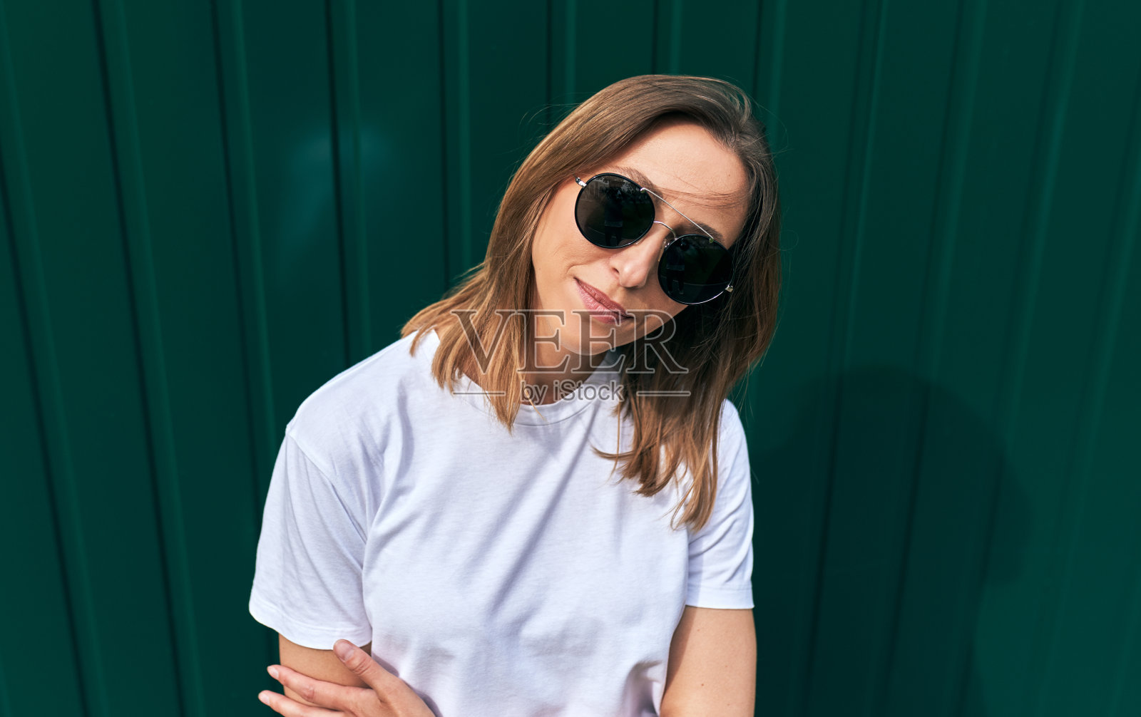 户外写真的欧洲年轻女子身穿白色t恤,戴着黑色太阳镜,望着镜头,背靠城市街道上的绿墙摆姿势。女学生微笑着走向学院。照片摄影图片