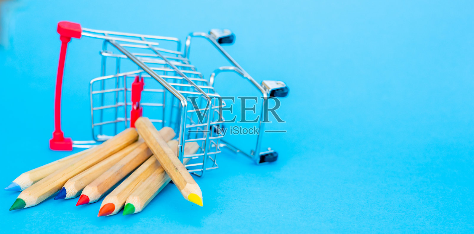 有一个购物篮的小手推车,用于在一个蓝色背景的商店购物。倒置的微型购物篮与分散的彩色铅笔。相机的焦点在铅笔上照片摄影图片