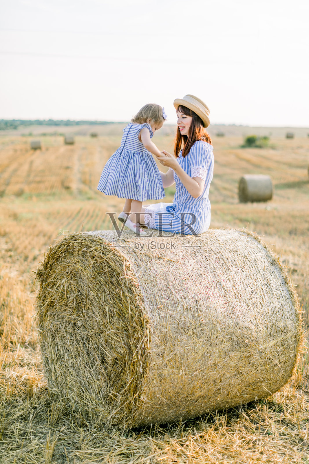 快乐可爱的小女孩孩子穿着蓝色条纹的裙子拥抱着她年轻的妈妈,一起坐在夏秋麦田里的干草圆捆上照片摄影图片